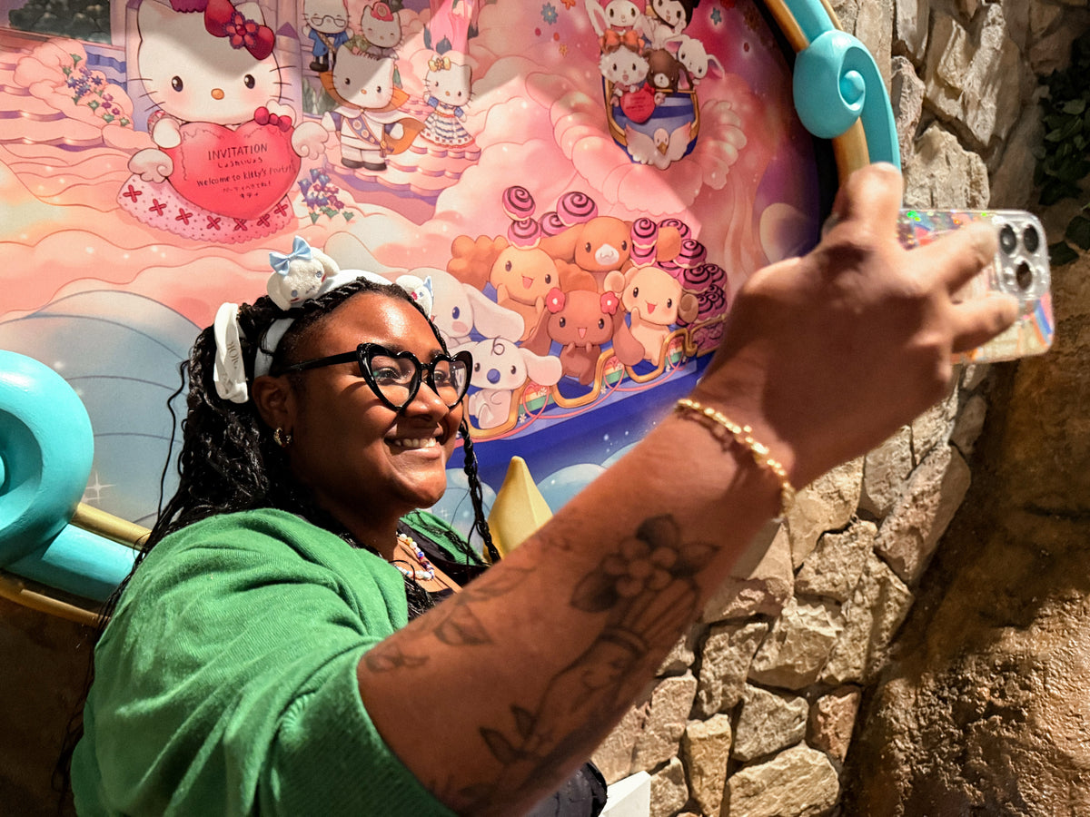 A woman wearing glasses and a green top takes a selfie in front of a colorful Hello Kitty and Sanrio character mural, with a stone wall visible on the right.