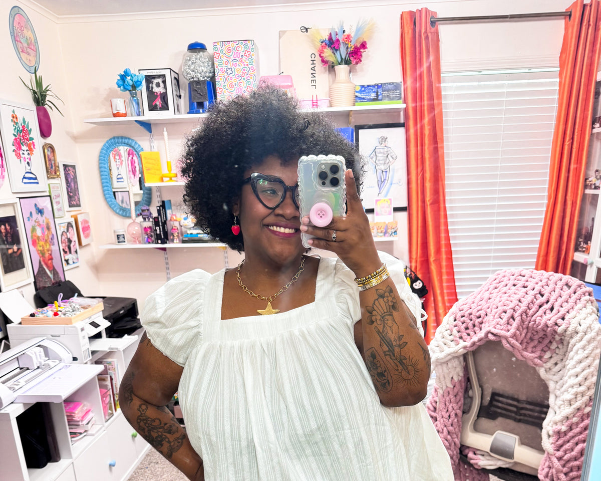 A woman takes a mirror selfie in a colorful, decorated room, wearing a white puff-sleeve top with a scalloped hem, dark jeans, and brown shoes.
